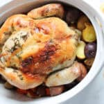 A roasted whole chicken with golden crispy skin sits in a white baking dish on a bed of assorted potatoes. Fresh herbs are on top, and lemon halves, butter, and a striped towel are beside the dish—perfect before learning how to clean a roasting pan.