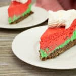 A slice of Christmas Peppermint Cheesecake with festive red, green, and brown layers, topped with a swirl of whipped cream, sits on a white plate. Another blurred slice rests in the background, both on a wooden surface.