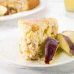 A slice of apple cake on a white plate, garnished with three fresh apple slices—perfect for St Patrick’s Day Desserts. In the background, a blurred plate with more cake and apple slices, and a glass of juice are visible.