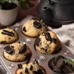A muffin tin filled with Oreo Muffins, each topped with Oreo pieces. Nearby, a small bowl of whole Oreos, a black teapot, a green plant in a white pot, and a light cloth complete the cozy scene.