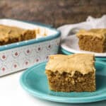A square slice of frosted Jif peanut butter cake sits on a turquoise plate in the foreground, with another slice on a white plate and the rest of the cake in a patterned baking dish in the background.