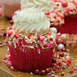 A cupcake in a red wrapper topped with white swirled frosting and colorful sprinkles, including pink, red, gold, and white candies, sits on a tray with more decorated cupcakes—perfect inspiration for Pink Cupcake Recipes.