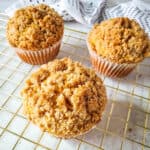 Three crumb-topped muffins sit on a gold cooling rack, highlighting the results of easy muffin recipes, with a white patterned cloth in the background on a light-colored surface.