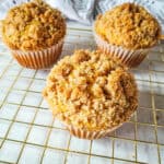 Three crumb-topped muffins sit on a gold cooling rack. With their golden, crumbly texture, they’re perfect for fall breakfast recipes, set against a light surface and a patterned cloth in the background.