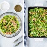 A bowl of Shredded Brussels Sprouts with Bacon sits next to a black baking dish filled with the same mixture; salt and pepper bowls and kitchen tongs are nearby on a white surface.