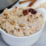 A white bowl filled with creamy oatmeal topped with chopped pecans sits on a gray cloth—an inviting Christmas breakfast idea, perfect for the crock pot—with raw oats and cinnamon sticks blurred in the background.