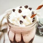 A glass of chocolate milkshake inspired by Starbucks Frozen Hot Chocolate, topped with whipped cream and chocolate chips, with a striped straw. A jar of chocolate chips and a striped cloth are in the background on a white surface.
