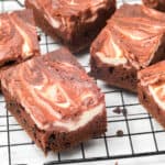 Six marbled red velvet brownies with swirls of chocolate and cream cheese are cooling on a black wire rack over a white surface. The brownies have a rich, fudgy texture and distinct marbled patterns on top.