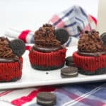 Three red velvet mini cheesecakes with chocolate frosting, chocolate chips, and mini Oreo cookies on top are arranged on a white plate—perfect as festive New Year’s Eve desserts. A checkered cloth and a glass of milk complete the scene.