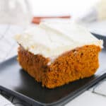 A square slice of moist pumpkin cake made with yellow cake mix and topped with creamy white frosting sits on a black plate atop a white checkered cloth. A blurred piping bag and plate are visible in the background.