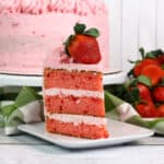 A slice of pink strawberry layer cake with pink frosting and a whole strawberry on top sits on a white plate—one of the sweetest birthday cakes ideas, with the rest of the cake and fresh strawberries in the background.