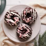 Three Mini Chocolate Peppermint Bundt Cakes topped with white icing and crushed peppermint, arranged on a light plate with a cream napkin, a glass of milk, and pine branches nearby.
