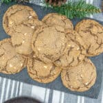 A slate serving tray with a stack of chocolate cookies featuring white chocolate chips, perfect for a Christmas Cookie Exchange, placed on a striped cloth with pinecones and greenery as festive decoration.