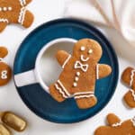 A gingerbread cookie decorated with white icing rests on the rim of a white cup on a blue plate, surrounded by more gingerbread desserts, gold measuring spoons, and a beige cloth napkin.