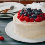 A frosted vanilla cake topped with fresh raspberries and blueberries sits on a white plate. Plates and forks are stacked in the background, with extra berries scattered on the table nearby—perfect for trying your favorite vanilla cake recipe.