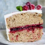 A slice of Cranberry Layer Cake with white frosting, red berry filling, and sugared cranberries on top, garnished with a mint leaf, sits on a white plate.