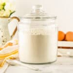 A large glass jar filled with homemade pancake mix sits on a white countertop, surrounded by a striped towel, white spoons, brown eggs in a tray, a glass bowl, and a vase of white flowers.