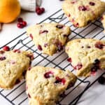 Triangular cranberry orange scones with a golden crust sit on a black cooling rack. Whole cranberries and an orange rest nearby on a white surface, adding a fresh look to the scene.
