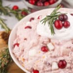 A bowl of cranberry fluff salad topped with whipped cream, fresh cranberries, and rosemary—perfect for Easy Thanksgiving Dessert Recipes—sits on a light table with scattered cranberries, rosemary, and a wooden spoon nearby.