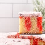 A close-up of a slice of Christmas poke cake with red gelatin swirls and pink whipped topping, lifted on a spatula above a pan of the same cake, against a white tiled background. Perfect inspiration for Christmas poke cake recipes.