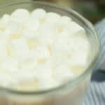 A close-up of a glass filled with hot chocolate topped with mini marshmallows, inspired by Starbucks drink recipes, with part of a blue and white striped cloth and a small yellow flower visible in the background.