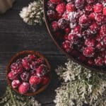 A large and small bowl of sugared cranberries sit on a dark wooden table, surrounded by frosted evergreen branches and a small bowl of sugar, capturing the festive charm of homemade sugared cranberries.