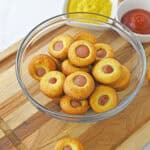A glass bowl filled with mini corn dog muffins sits on a wooden cutting board, making perfect easy appetizers for a party, with small bowls of mustard and ketchup in the background.