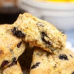 A close-up of golden-brown blueberry scones stacked on a white plate, with one scone broken in half to reveal the moist interior and visible blueberries—perfect for fans of lemon desserts. A blurred bowl appears in the background.