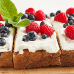 Three slices of cake—made using pancake mix—are topped with whipped cream, fresh raspberries, blueberries, and a sprig of mint, artfully arranged on a wooden surface.