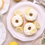 Three mini lemon bundt cakes with white icing and lemon slices are served on a white plate, surrounded by extra plates, forks, whole lemons, and white flowers artfully arranged on a light surface.