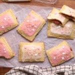 Six rectangular pastries with pink and white icing and heart-shaped sprinkles rest on parchment paper; one reveals a red filling. A dish towel, pink glaze, and sprinkles are nearby—perfect for your list of cozy winter snack ideas.