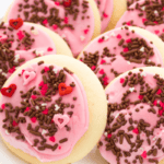 A plate of sugar cookies inspired by the classic Lofthouse sugar cookie recipe, topped with pink frosting and decorated with chocolate sprinkles and small red and pink heart-shaped sprinkles.