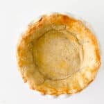 A golden-brown, baked pie crust sits in a white pie dish on a plain white background, showing the perfect result of how to make a pie crust with no filling inside.
