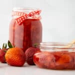 Two glass jars filled with strawberry preserves, one tied with a red checkered ribbon, sit beside fresh strawberries. A wooden spoon rests in the open jar, all arranged on a white surface with a plain background.