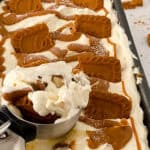 A close-up of a tray of creamy Biscoff Cheesecake Ice Cream topped with caramel sauce and Biscoff cookies, with an ice cream scoop lifting a serving. Crumbled cookies are scattered around the tray.