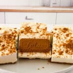 Four pieces of creamy Biscoff Cheesecake Bars topped with crumbled Lotus Biscoff cookies and a whole cookie in the center, displayed on a white plate in a modern kitchen setting.