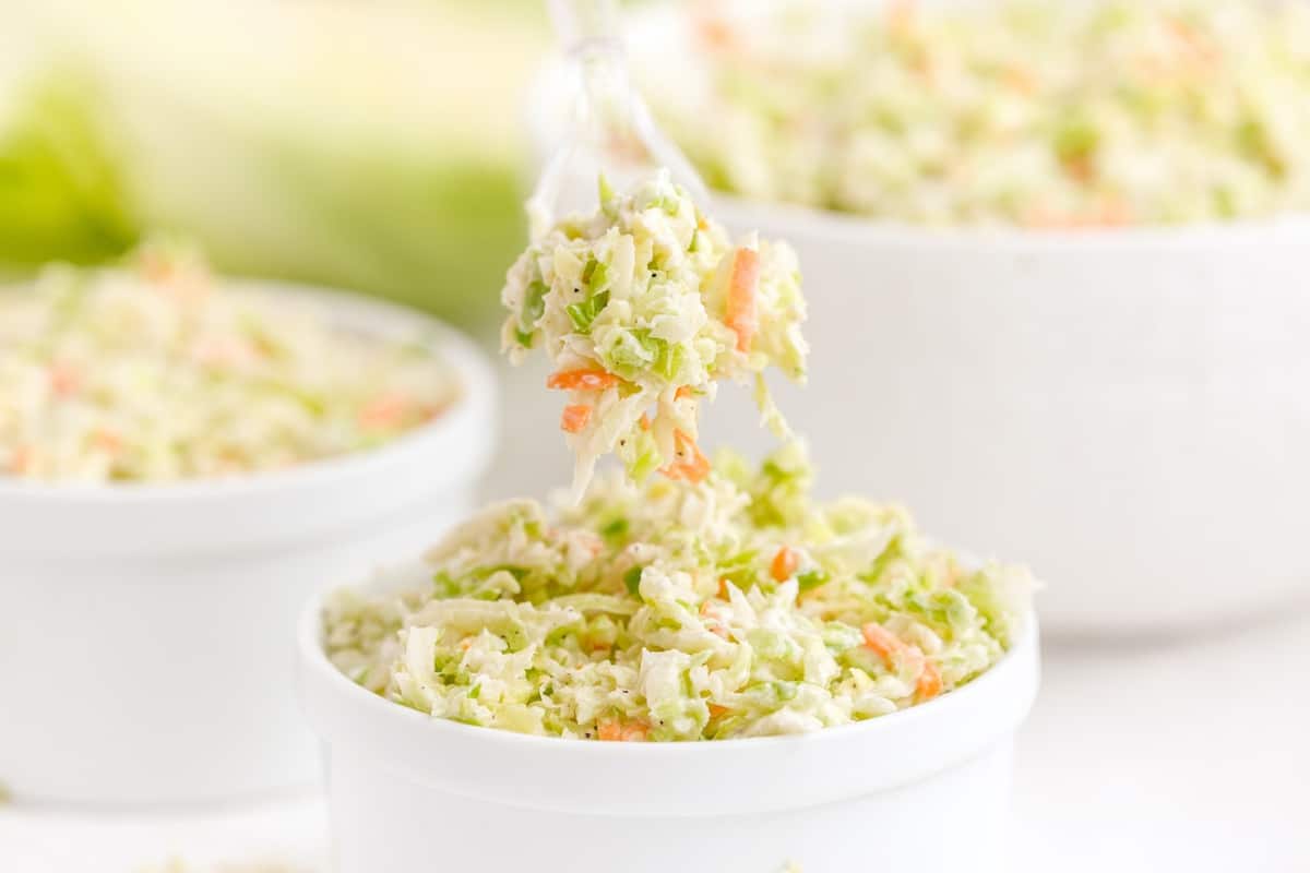 A close-up of creamy coleslaw in white bowls, with a fork lifting a portion. The coleslaw contains shredded cabbage, carrots, and dressing. The background is softly blurred.