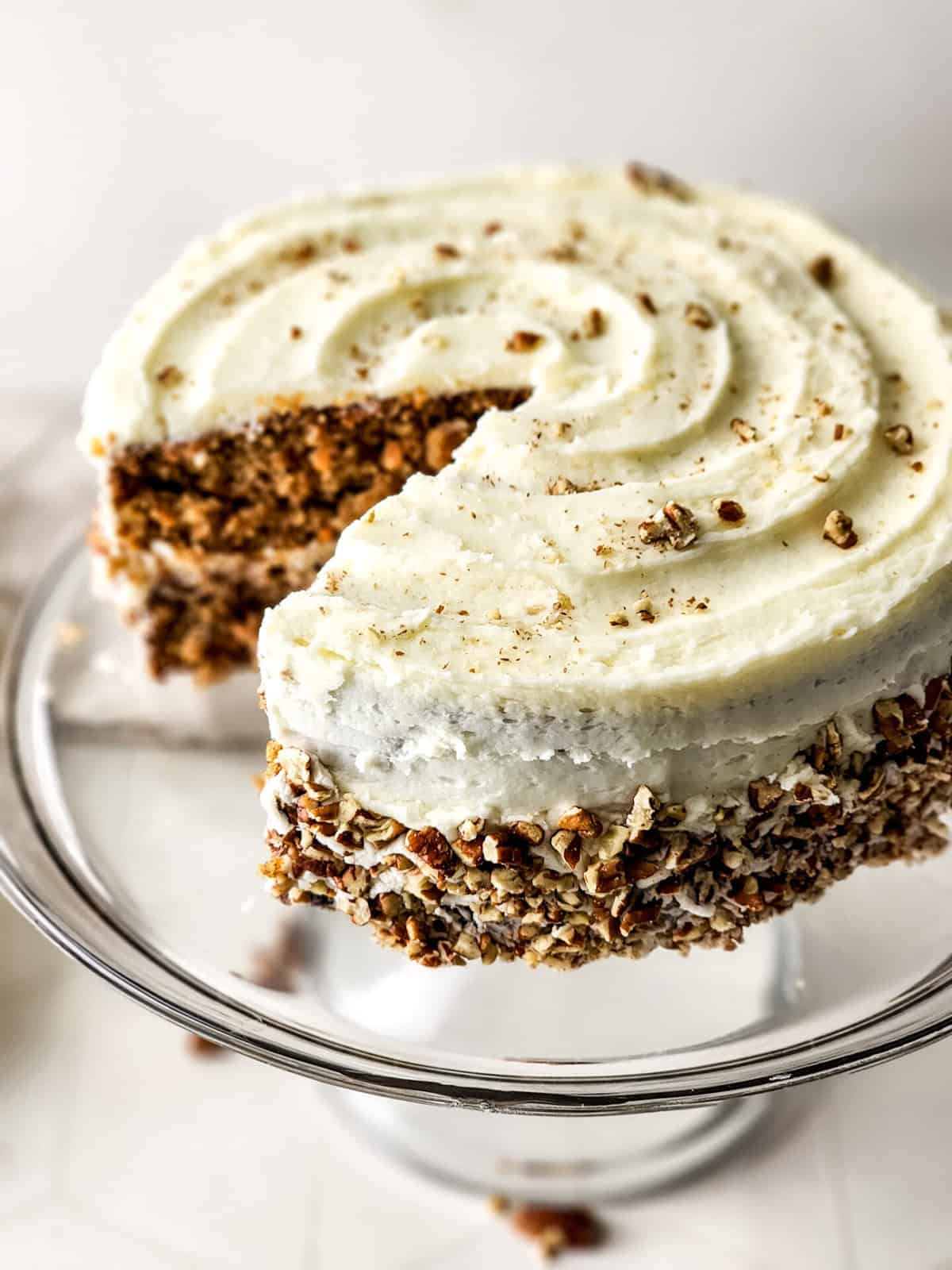 A frosted layer cake made from the best carrot cake recipe sits on a glass stand, its sides covered in chopped nuts. A large slice reveals a moist, textured interior, while swirled white frosting on top is sprinkled with more nuts.