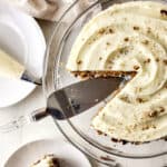 A glass cake stand holds a round cake with creamy white frosting and sprinkled nuts, made from the best carrot cake recipe. A slice is being lifted with a cake server. Nearby are a piping bag, a plate, and a beige napkin.