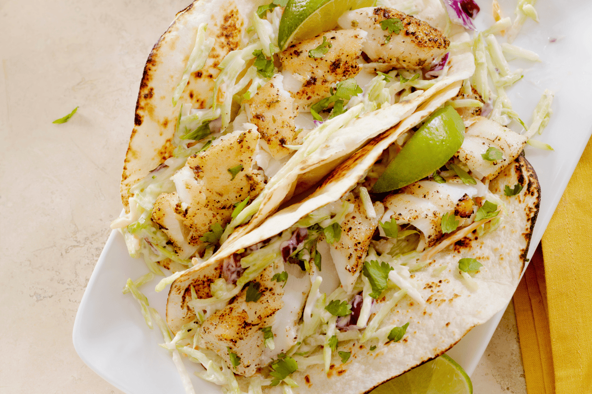 Two grilled fish for fish tacos topped with shredded cabbage, cilantro, and creamy sauce are served on a white plate with lime wedges.