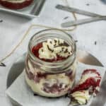 A Red Velvet Cake Jar filled with layers of rich cake and cream cheese frosting, topped with chocolate sprinkles, sits on a plate beside a spoon with a bite of cake.