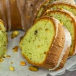 Close-up of sliced pistachio bundt cake, made with yellow cake mix and topped with glaze, revealing a green interior and sprinkled pistachio pieces on a parchment-lined baking tray. Great for creative uses for yellow cake mix.