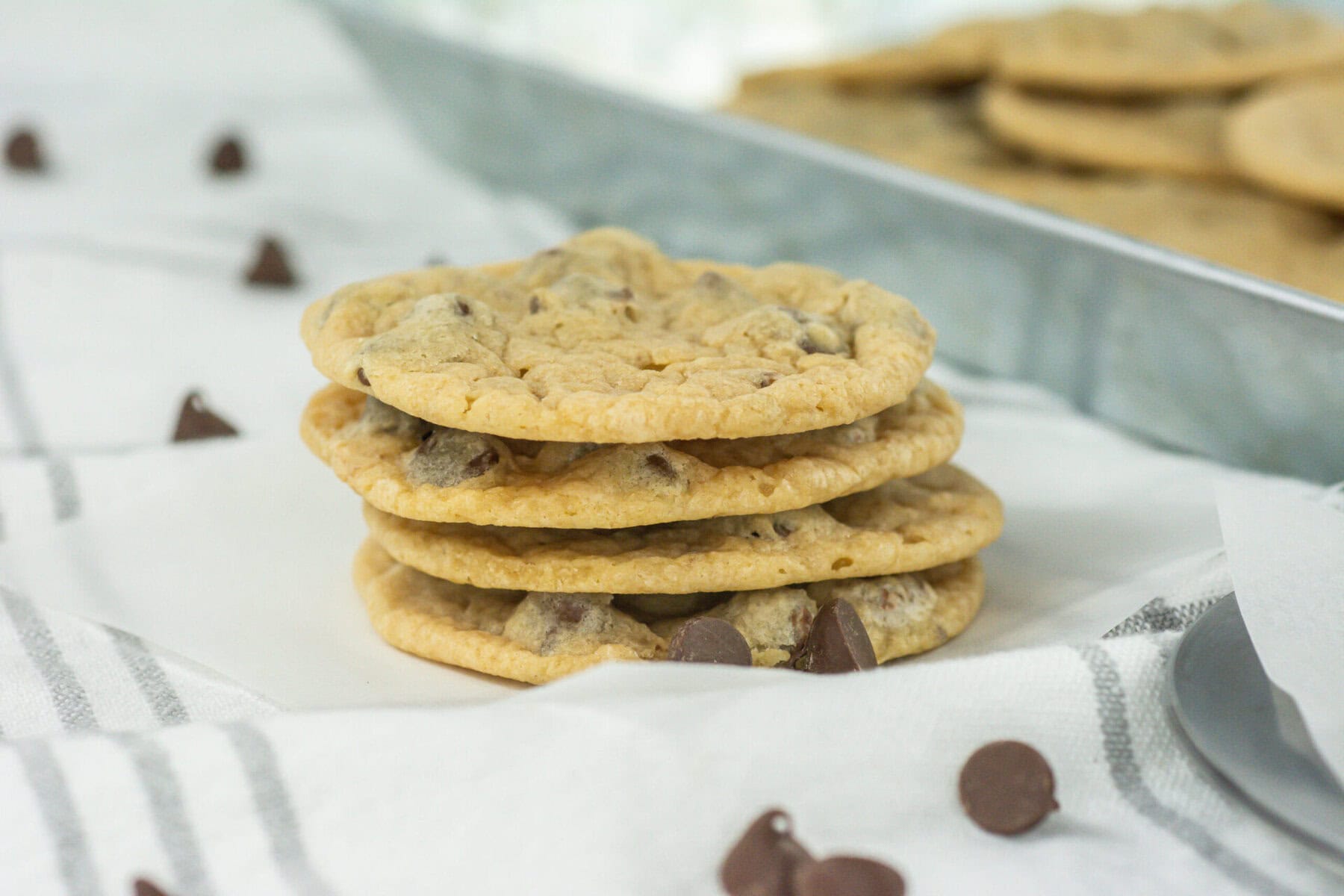 A stack of four chocolate chip cookies sits on a white cloth napkin with chocolate chips scattered around and more cookies on a tray in the background.
