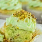 A close-up of a pistachio cupcake with light green frosting and chopped pistachios on top—an ideal St Patrick’s Day Dessert. The cupcake is partially unwrapped and cut in half, revealing its soft, green interior. Other cupcakes are blurred in the background.