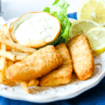 A plate with beer battered fish fillets, French fries, lemon slices, lettuce, and a small bowl of tartar sauce, set on a blue napkin.