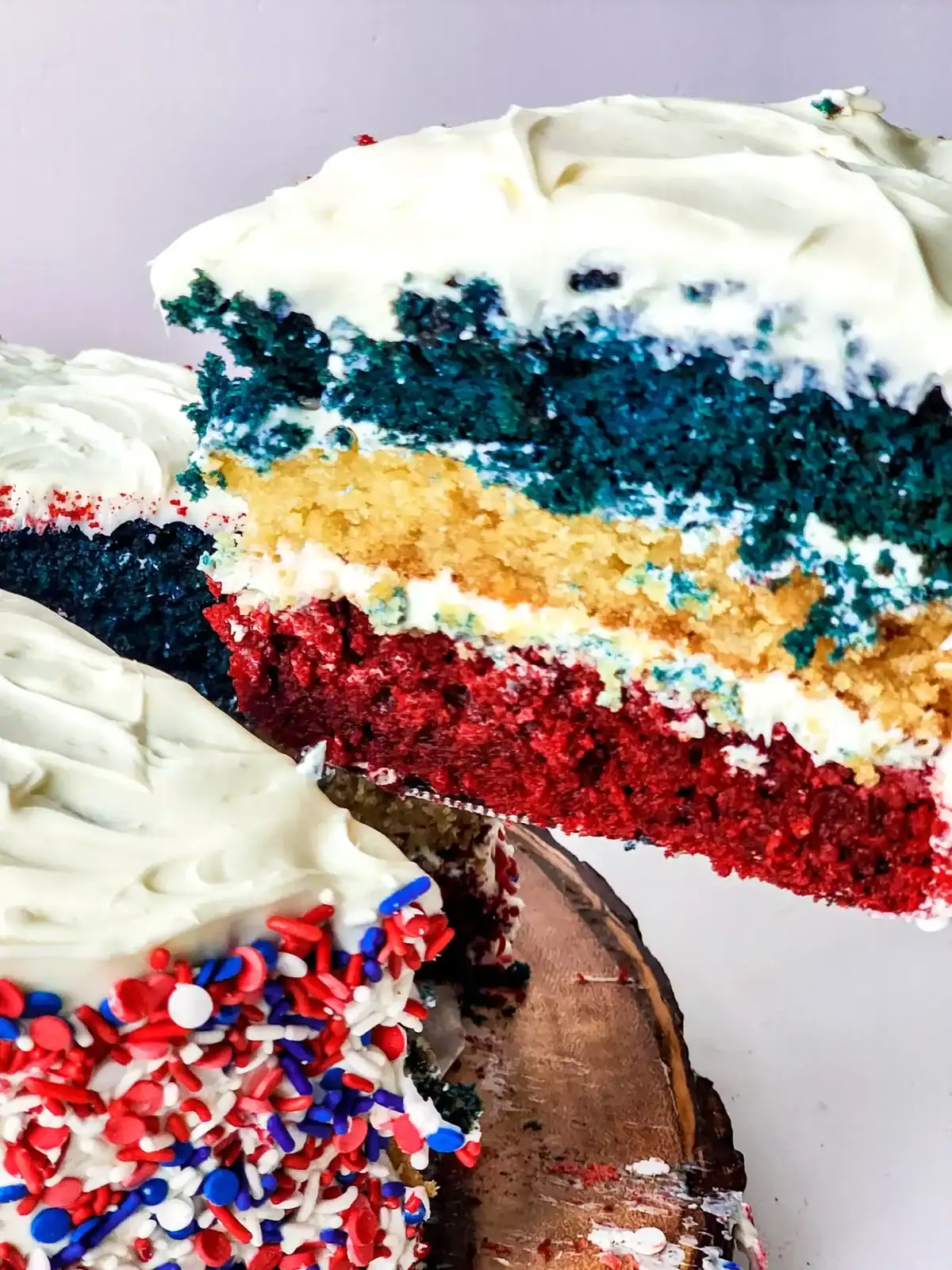 A slice of layered cake with red, white, and blue layers and white frosting is being lifted from the whole cake, which is decorated with red, white, and blue sprinkles—perfect for Memorial Day desserts.