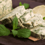 Slices of herb butter with chopped green herbs are arranged on a dark plate, garnished with fresh mint leaves and lime slices in the background—perfect for learning how to make herb butter with dried herbs.
