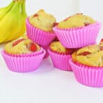 Six strawberry banana muffins in pink paper liners are stacked on a white surface. In the background, fresh bananas and a few strawberries add to the brightly lit, colorful scene.