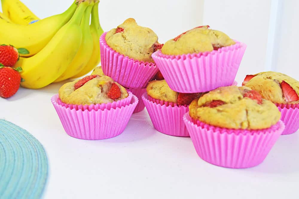 Six strawberry banana muffins in pink paper liners are stacked on a white surface. In the background, fresh bananas and a few strawberries add to the brightly lit, colorful scene.
