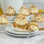 Cupcakes with swirled cinnamon frosting are arranged on a counter. One cupcake is cut open on a stack of plates, showing its fluffy inside. A fork rests nearby, and all cupcakes are in white paper liners.